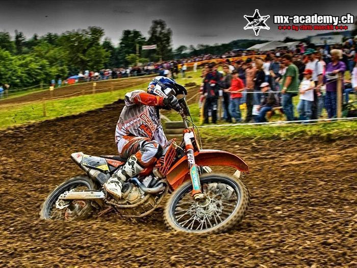 Motocross Rennen: Fahrer in der Kurve, dahinter verfolgen viele Zuschauer das Geschehen am Streckenrand