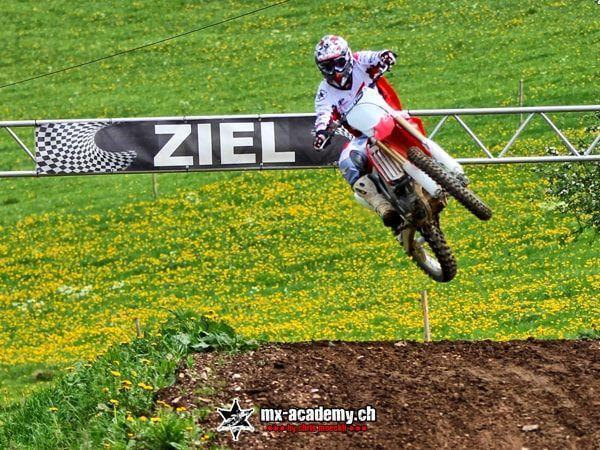 Motorsport Veranstaltung: Fahrer springt vor einem Ziel-Banner über die Wiese voller Löwenzahn