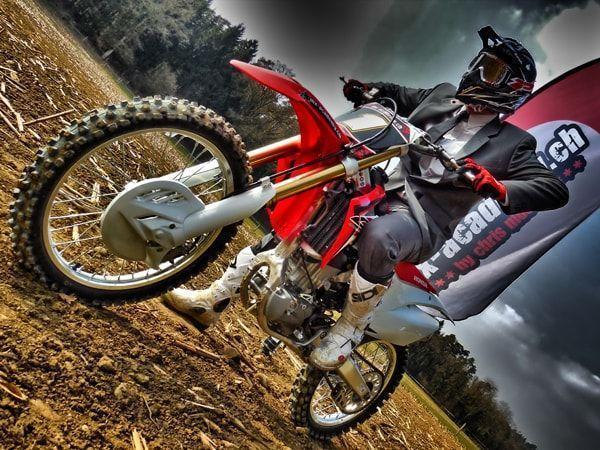 Betriebsausflug mit Motocross: Fahrer im Anzug mit Krawatte auf einer roten Honda, daneben eine Fahne