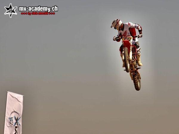 Motocross Fahrer im Sprung vor grauem Himmel, unten links eine Fahne – Rennen und Begriffe im MX Sport