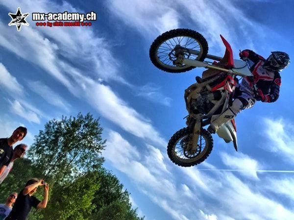Fahrer im Sprung, von unten fotografiert, links schauen Zuschauer zu – Szene wie bei Motocross Rennen