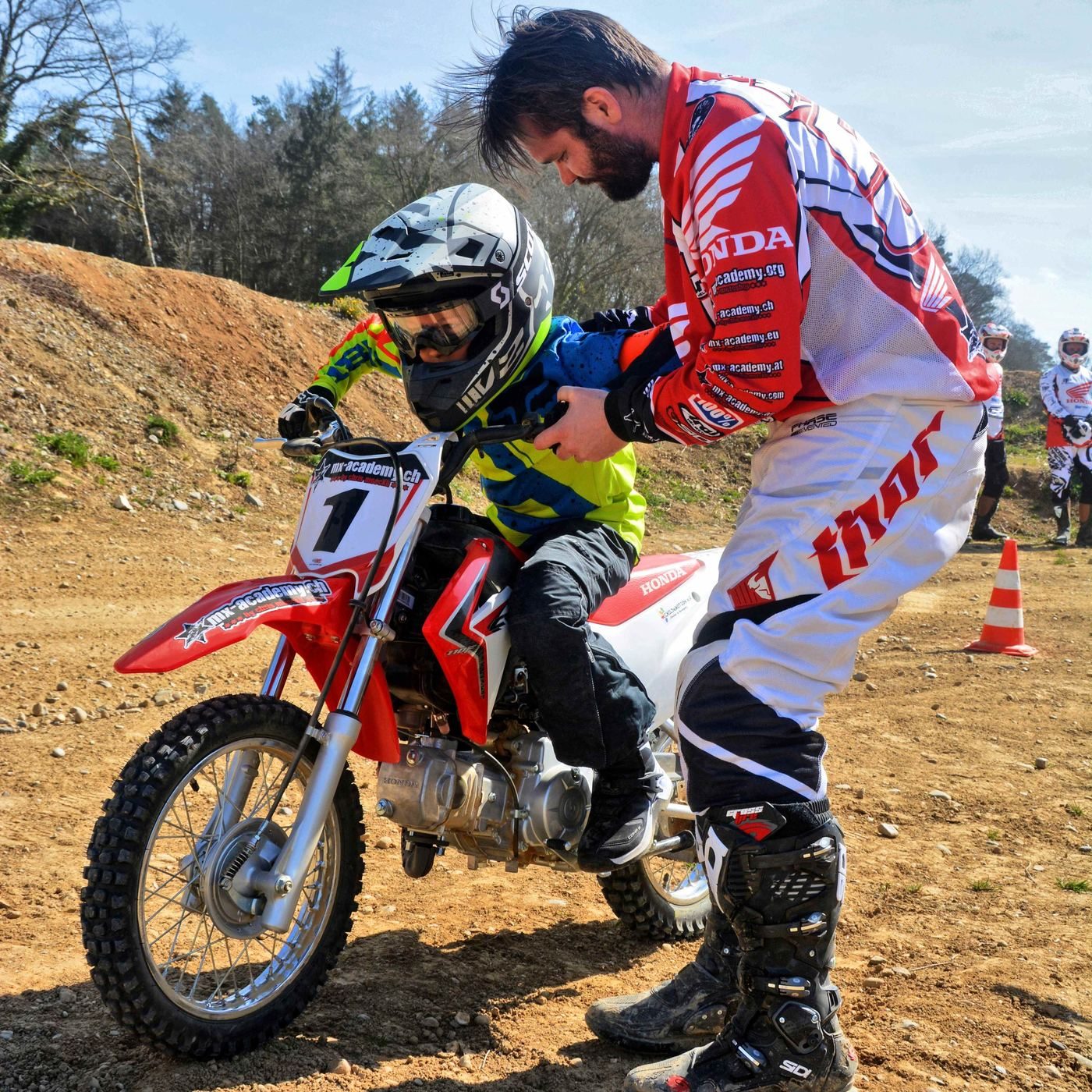 Trainer kniet neben einem Kind auf seinem Kindermotorrad und erklärt – Kindercross mit persönlicher Betreuung